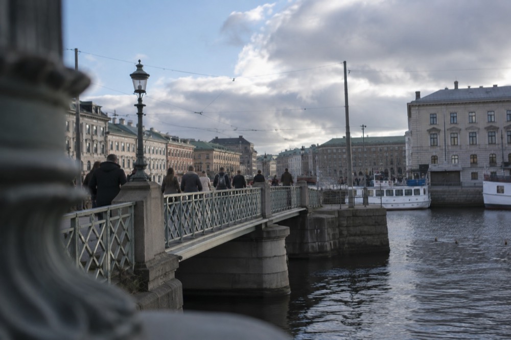 Pedestrians crossing a canal bridge in Gothenburg with moored boats and brick buildings behind them