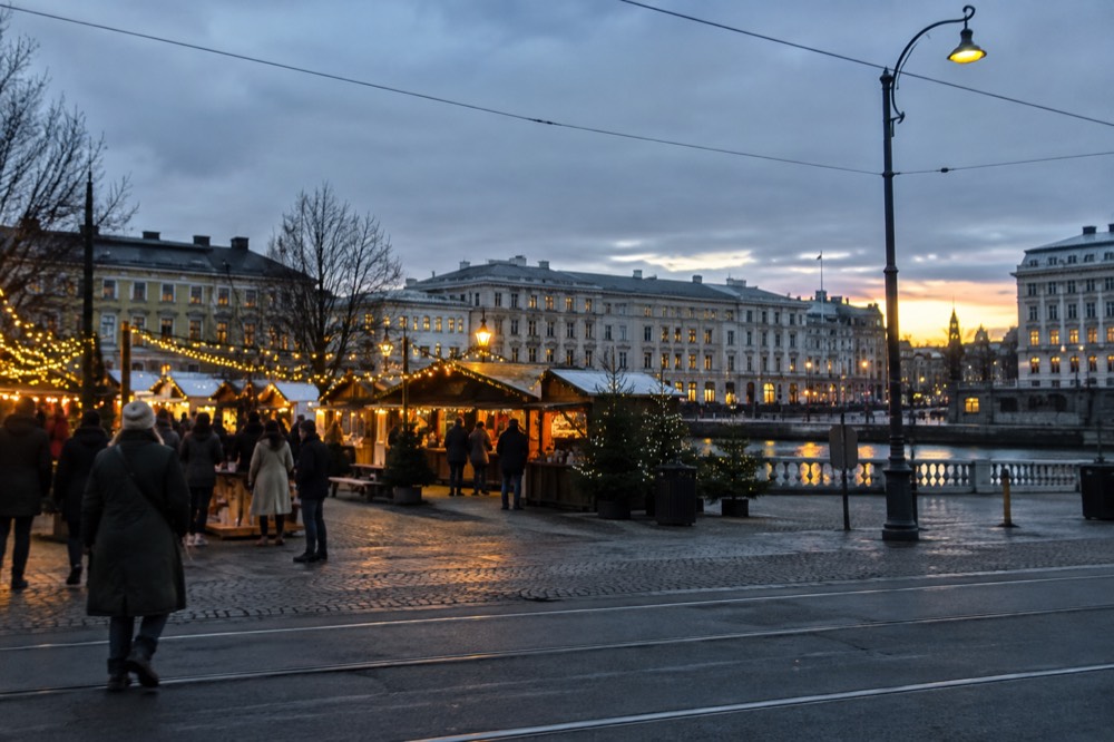 Festive market stalls and string lights in a Scandinavian city square at dusk, with people in winter coats browsing under a dark blue sky