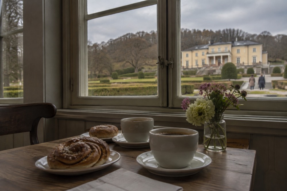 A café table with coffee, cinnamon buns and a small vase of flowers beside a window in a wooden building