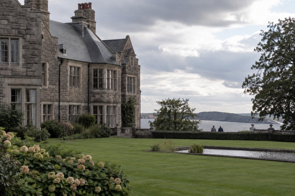 A wooden country manor with pale facades seen across a formal garden lawn