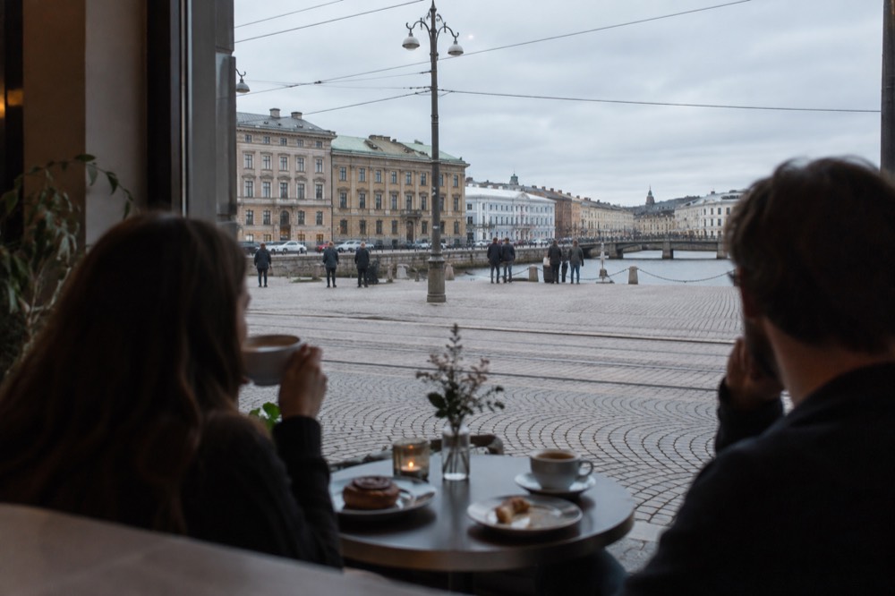 Two people sitting at a cafe window table with coffee cups and pastries in a Gothenburg cafe