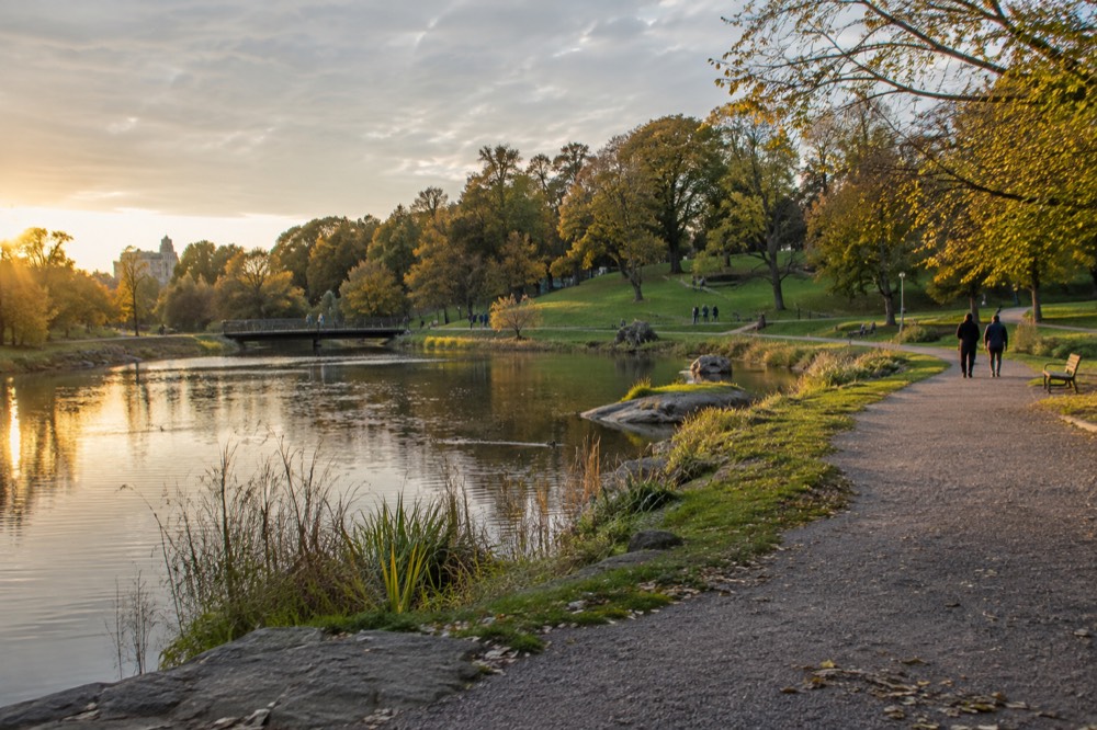 A green park hillside in Gothenburg with a pond, trees and people walking on gravel paths