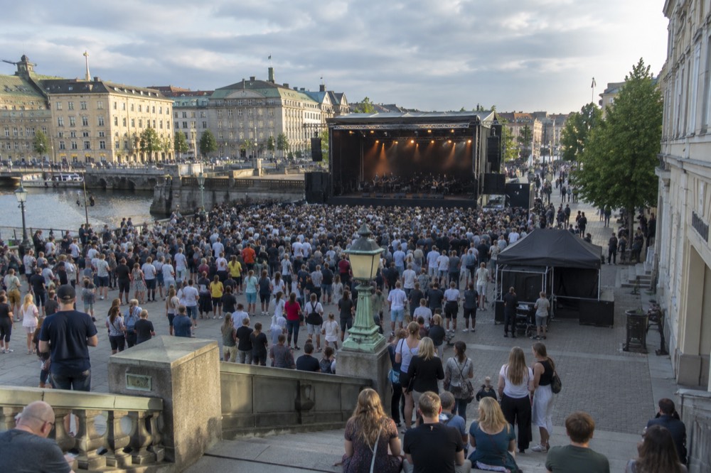 An outdoor concert stage on a wide city avenue lined with stone buildings and crowds in summer evening light