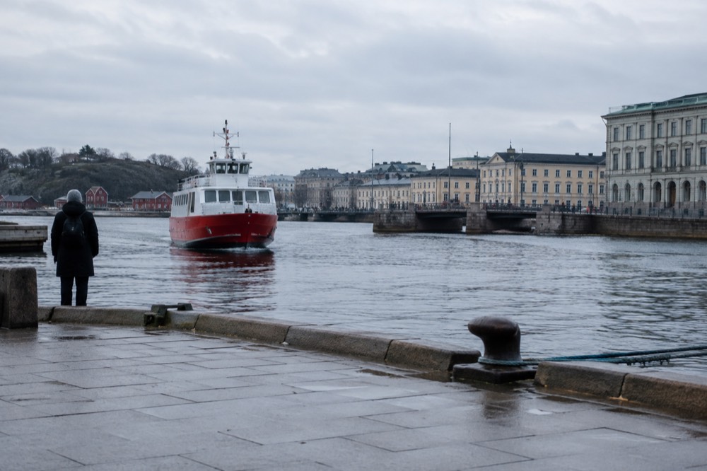 A red ferry approaching a rocky island quay with small wooden boathouses